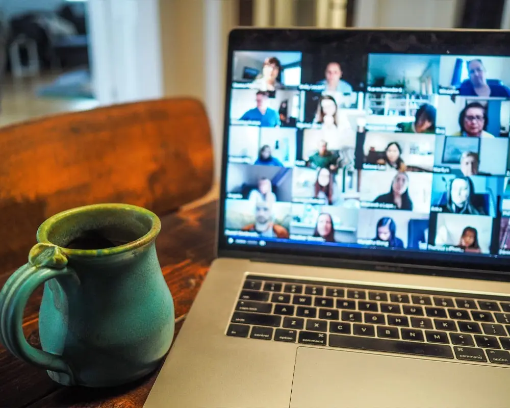 A Zoom call displayed on a laptop screen with multiple participants visible, accompanied by a steaming coffee mug placed nearby, highlighting productivity, virtual teamwork, and casual workplace collaboration.