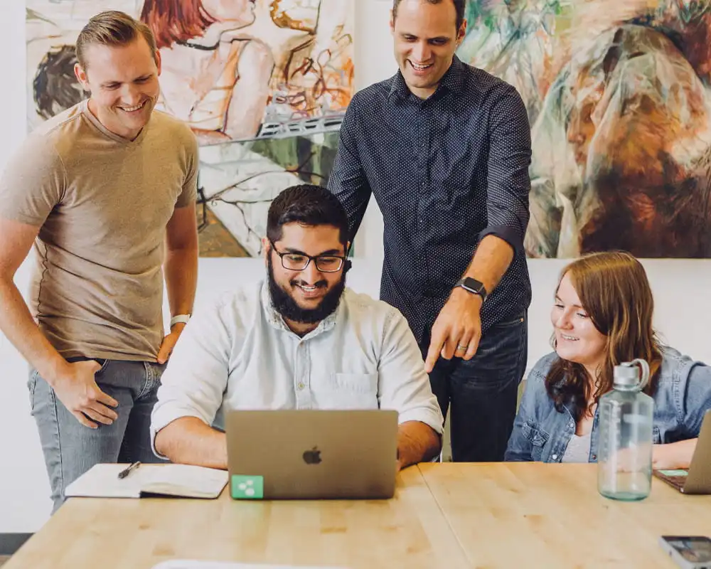 Three men and one woman dressed professionally, gathered around a MacBook, smiling and gesturing toward the screen in an engaging moment of collaboration, teamwork, and shared enthusiasm.