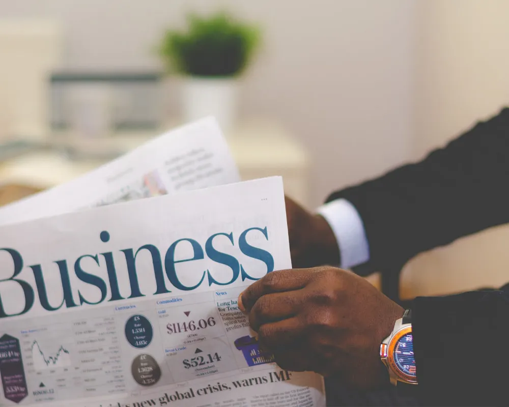 An African American individual dressed in professional attire, confidently holding a business newspaper open to financial reports, reflecting focus, knowledge, and engagement with economic trends.